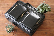Black leather backpack briefcase Elegant gold zipper detailing Luxury business accessory Wooden table setting Two frontal zip compartments Signature script logo on front High-quality professional bag Versatile briefcase with backpack option Puffy leather texture Sleek modern design Stylish workplace gear